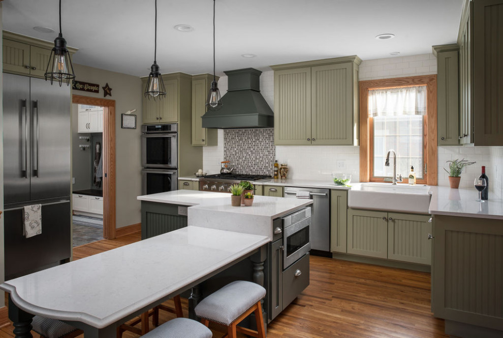 This is an example of a large traditional l-shaped eat-in kitchen in Columbus with a farmhouse sink, recessed-panel cabinets, green cabinets, quartz benchtops, white splashback, ceramic splashback, stainless steel appliances, medium hardwood floors, multiple islands, brown floor and white benchtop.