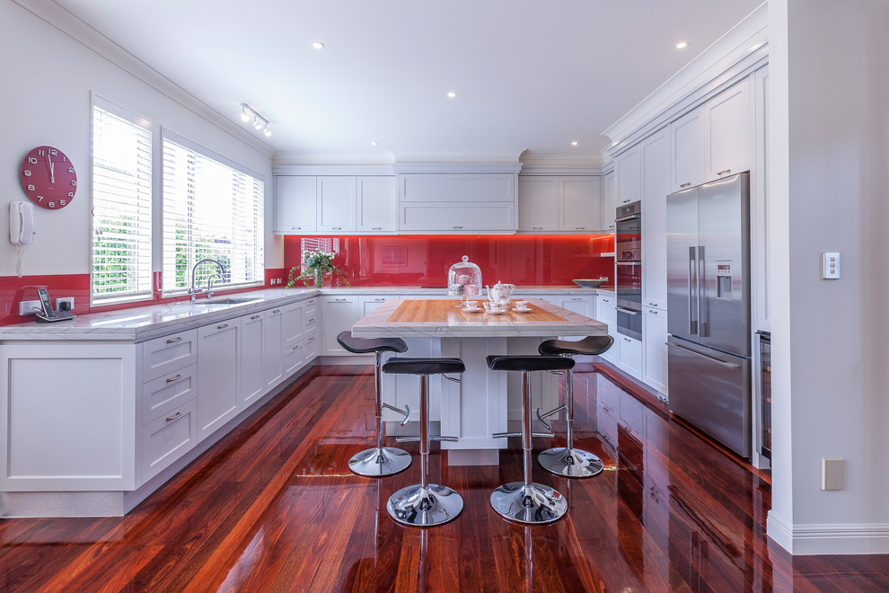 Photo of a large traditional u-shaped eat-in kitchen in Auckland with an undermount sink, recessed-panel cabinets, granite benchtops, red splashback, glass sheet splashback, stainless steel appliances, dark hardwood floors, with island, brown floor and white cabinets.