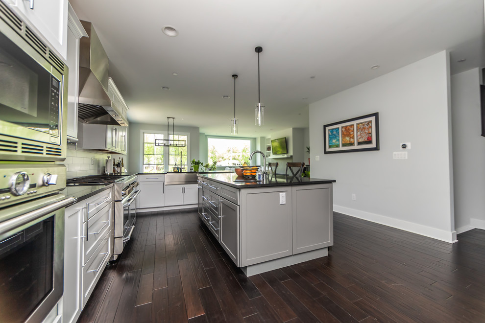 Large contemporary u-shaped open plan kitchen in Baltimore with a farmhouse sink, shaker cabinets, white cabinets, white splashback, stainless steel appliances, dark hardwood floors, with island, brown floor, solid surface benchtops, ceramic splashback and black benchtop.