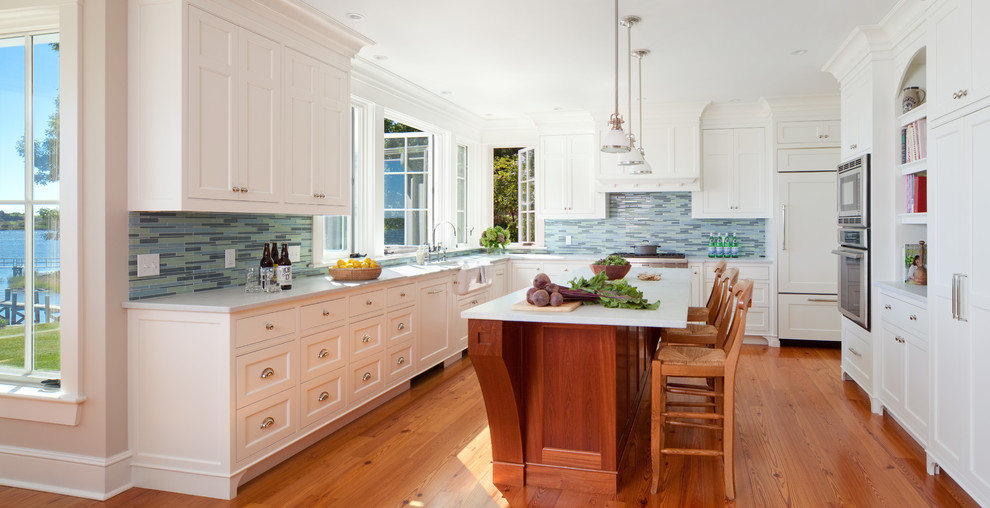 Design ideas for a beach style kitchen in Providence with a farmhouse sink, recessed-panel cabinets, white cabinets, marble benchtops, blue splashback, glass tile splashback, panelled appliances, medium hardwood floors, with island, brown floor and white benchtop.