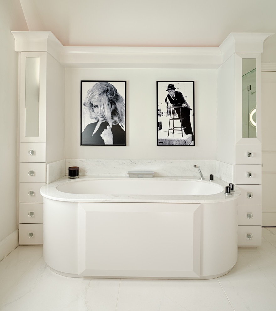 This is an example of a contemporary bathroom in London with flat-panel cabinets, white cabinets, marble benchtops, white walls, marble floors and a drop-in tub.