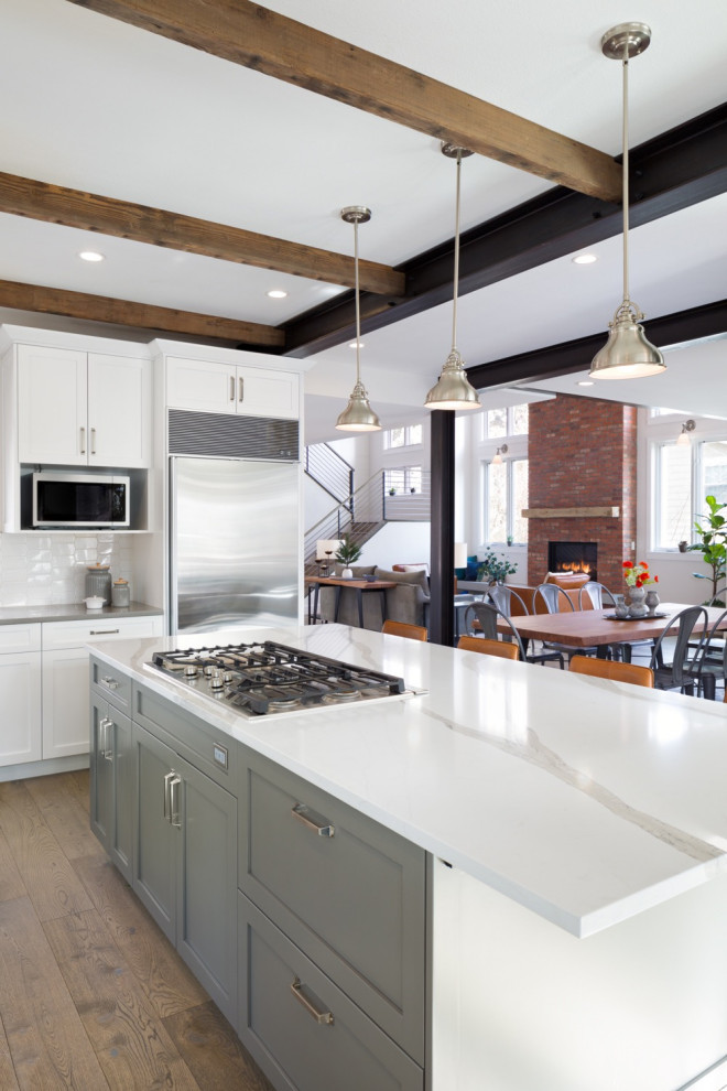 Photo of a large industrial l-shaped eat-in kitchen in Denver with a drop-in sink, recessed-panel cabinets, white cabinets, marble benchtops, white splashback, ceramic splashback, stainless steel appliances, medium hardwood floors, with island, beige floor and grey benchtop.