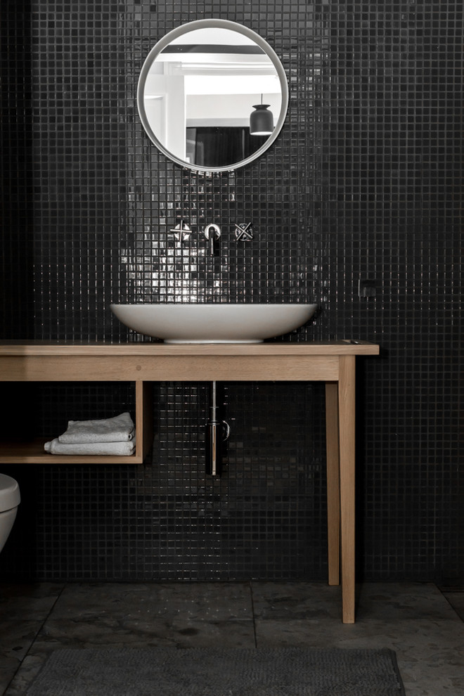 Mid-sized modern powder room in Berlin with furniture-like cabinets, medium wood cabinets, a wall-mount toilet, black tile, mosaic tile, black walls, porcelain floors, a vessel sink, wood benchtops and beige benchtops.