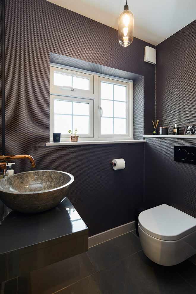 Photo of a contemporary powder room in London with a wall-mount toilet, gray tile, a vessel sink, black floor and grey benchtops.