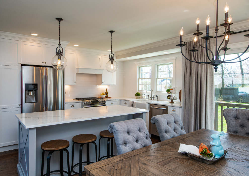Mid-sized transitional l-shaped eat-in kitchen in New York with a farmhouse sink, shaker cabinets, white cabinets, quartz benchtops, white splashback, subway tile splashback, stainless steel appliances, dark hardwood floors, with island, brown floor and white benchtop.