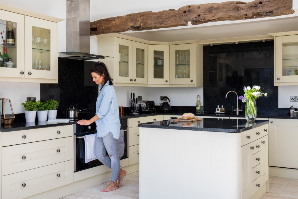 Large traditional cream and black l-shaped open plan kitchen in West Midlands with a double-bowl sink, glass-front cabinets, white cabinets, granite worktops, black splashback, laminate floors, an island, black worktops and exposed beams.