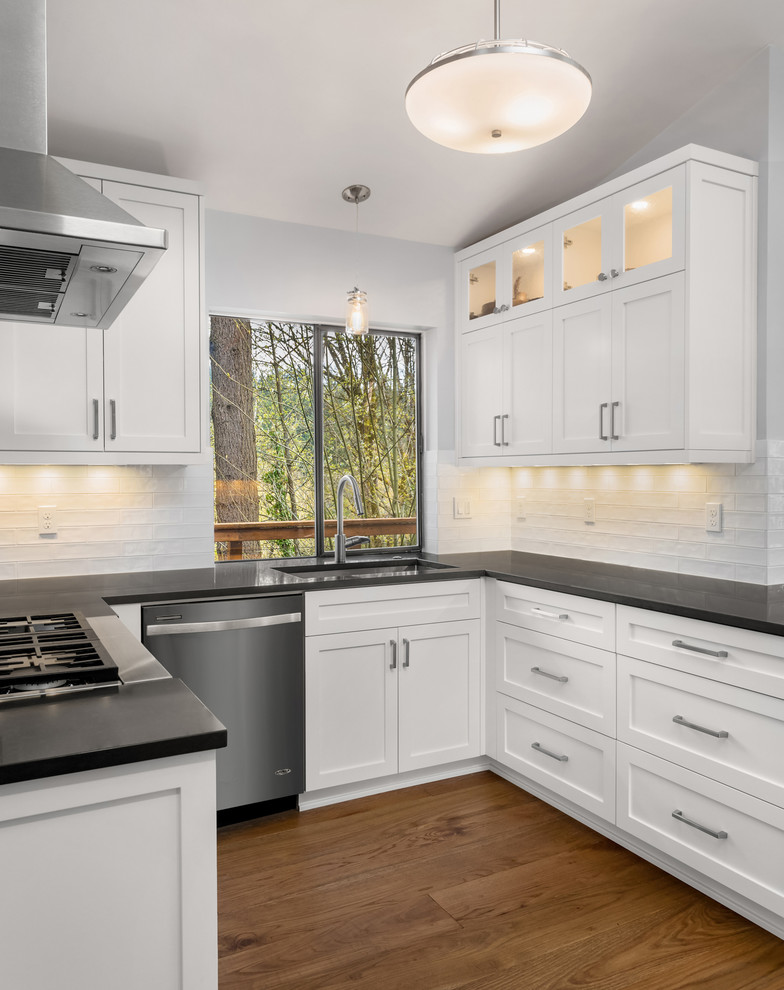 This is an example of a transitional u-shaped kitchen in Portland with an undermount sink, shaker cabinets, white cabinets, white splashback, subway tile splashback, stainless steel appliances, medium hardwood floors, brown floor and black benchtop.