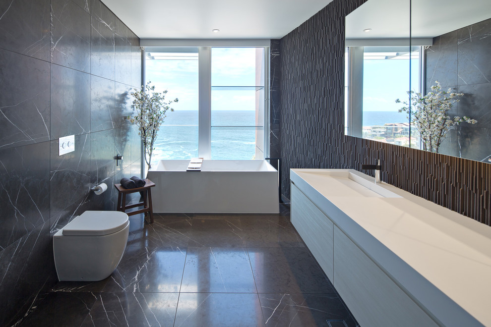 This is an example of a contemporary master bathroom in Sydney with light wood cabinets, a freestanding tub, brown tile, stone tile, marble floors, engineered quartz benchtops, flat-panel cabinets, a wall-mount toilet and an integrated sink.
