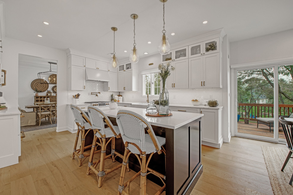 This is an example of a large transitional u-shaped eat-in kitchen in Philadelphia with a farmhouse sink, recessed-panel cabinets, white cabinets, quartz benchtops, white splashback, subway tile splashback, white appliances, light hardwood floors, with island, brown floor and white benchtop.
