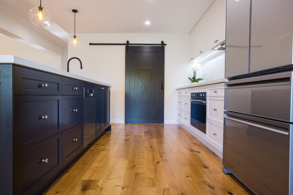 This is an example of an expansive contemporary galley kitchen pantry in Brisbane with a double-bowl sink, shaker cabinets, black cabinets, quartz benchtops, grey splashback, subway tile splashback, stainless steel appliances, medium hardwood floors, with island, brown floor and white benchtop.