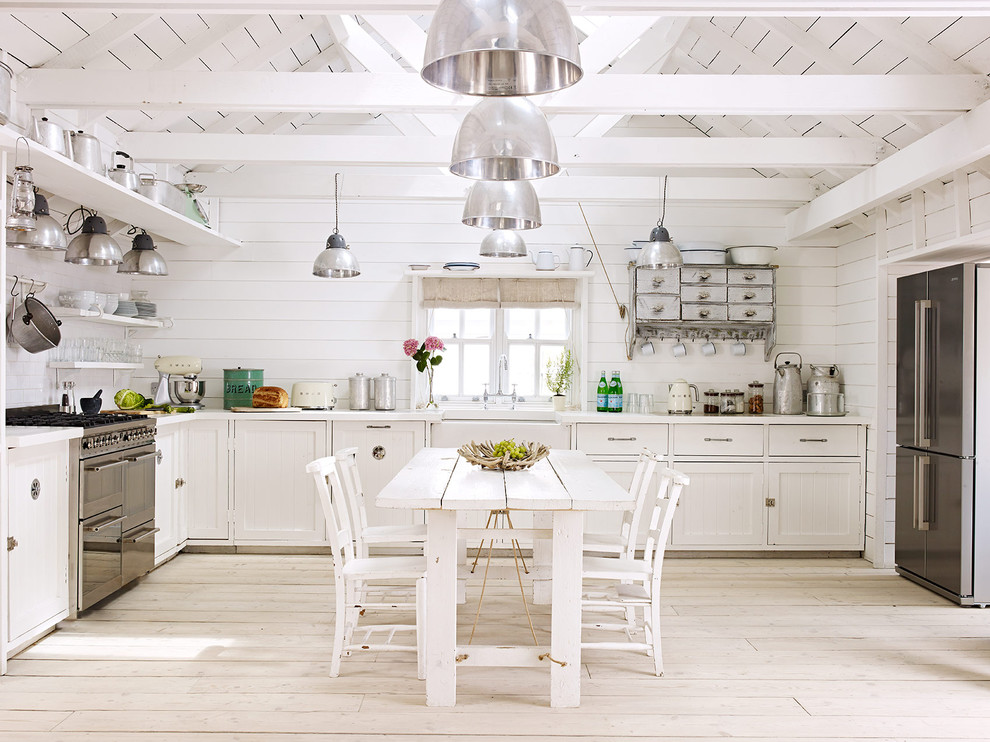 Photo of a rural u-shaped kitchen/diner with a belfast sink, shaker cabinets, white cabinets, white splashback, stainless steel appliances and light hardwood flooring.