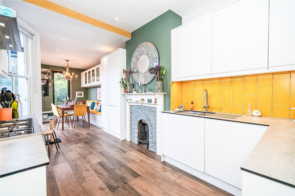 This is an example of a small contemporary kitchen/diner in London with a submerged sink, flat-panel cabinets, white cabinets, composite countertops, yellow splashback, porcelain splashback, stainless steel appliances, porcelain flooring, no island, brown floors, grey worktops and a chimney breast.