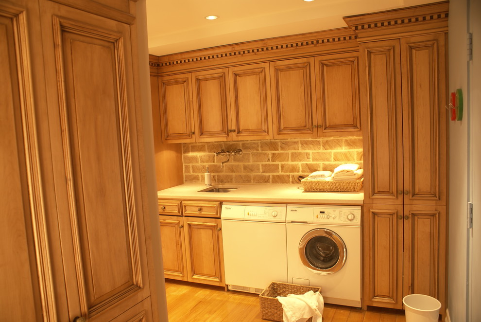 This is an example of a small traditional l-shaped laundry room in New York with a single-bowl sink, raised-panel cabinets, marble benchtops, yellow walls, light hardwood floors, a side-by-side washer and dryer and medium wood cabinets.