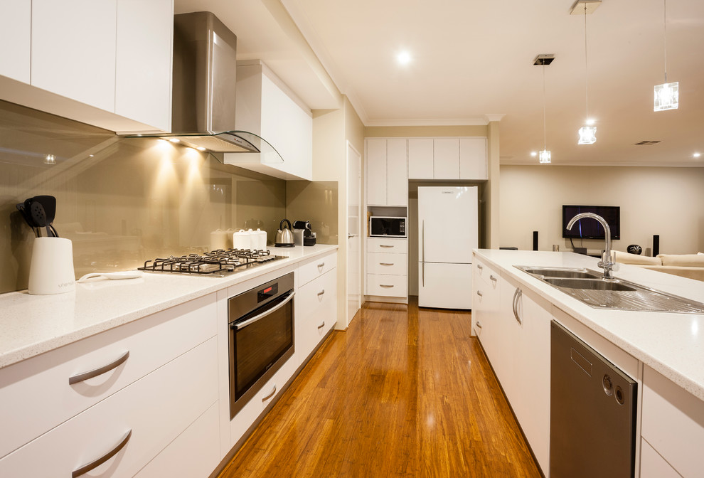 Photo of a contemporary galley open plan kitchen in Perth with a double-bowl sink, flat-panel cabinets, white cabinets, beige splashback, glass sheet splashback, stainless steel appliances and medium hardwood floors.