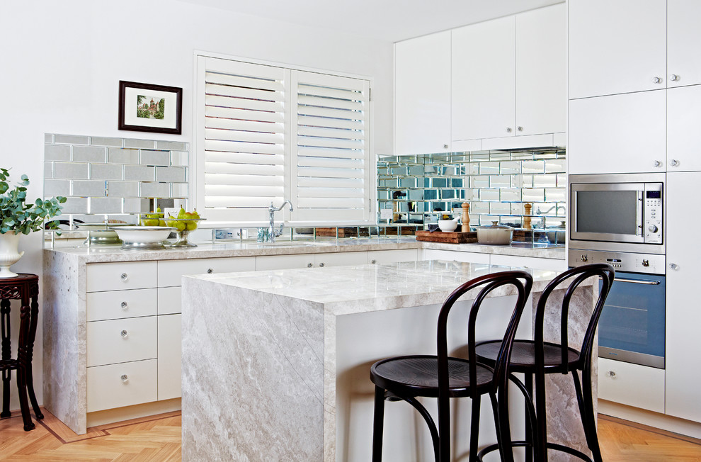 This is an example of a transitional l-shaped kitchen in Sydney with an undermount sink, flat-panel cabinets, white cabinets, metallic splashback, subway tile splashback, stainless steel appliances, light hardwood floors and with island.