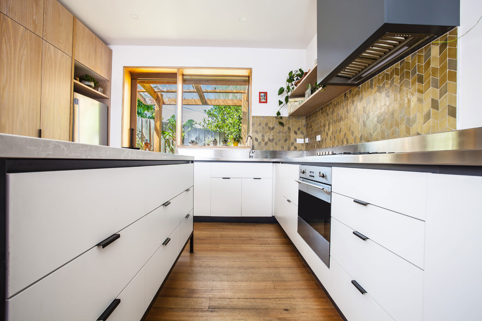 This is an example of a mid-sized contemporary u-shaped open plan kitchen in Melbourne with a double-bowl sink, flat-panel cabinets, white cabinets, stainless steel benchtops, green splashback, ceramic splashback, stainless steel appliances, medium hardwood floors and with island.