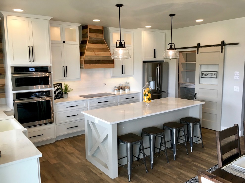 This is an example of a mid-sized country u-shaped eat-in kitchen in Other with a farmhouse sink, shaker cabinets, white cabinets, marble benchtops, white splashback, subway tile splashback, stainless steel appliances, vinyl floors, with island and brown floor.