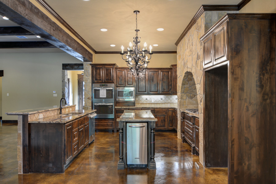 Example of a mid-sized mountain style concrete floor eat-in kitchen design with an undermount sink, raised-panel cabinets, dark wood cabinets, granite countertops, beige backsplash, stone tile backsplash, stainless steel appliances and an island
