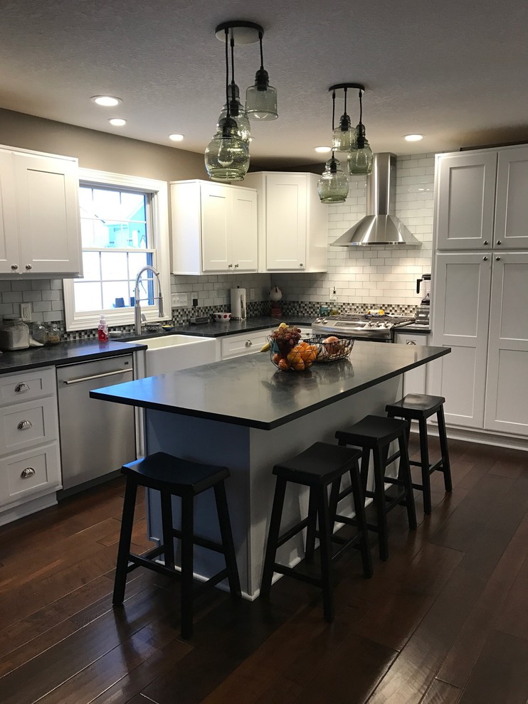 This is an example of a mid-sized contemporary l-shaped eat-in kitchen in Cedar Rapids with a farmhouse sink, flat-panel cabinets, white cabinets, soapstone benchtops, white splashback, glass tile splashback, stainless steel appliances, medium hardwood floors, with island, brown floor and grey benchtop.