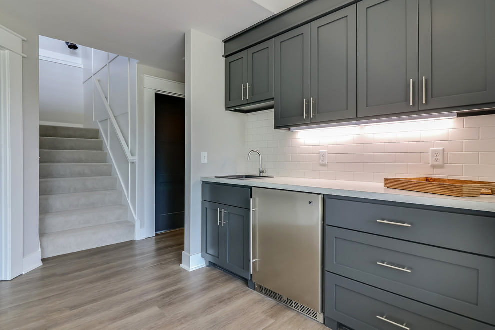 This is an example of a mid-sized contemporary single-wall wet bar in Grand Rapids with grey cabinets, white splashback, subway tile splashback, light hardwood floors, beige floor, grey benchtop and a drop-in sink.
