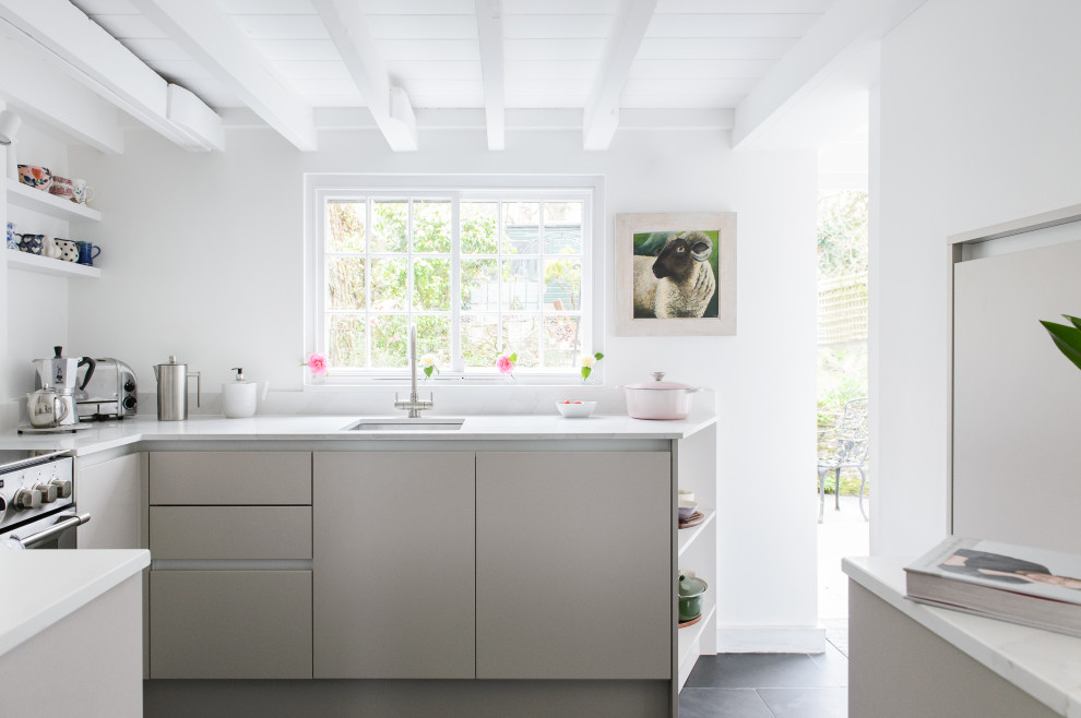 Small contemporary grey and cream u-shaped enclosed kitchen in Cornwall with a built-in sink, flat-panel cabinets, beige cabinets, quartz worktops, white splashback, engineered quartz splashback, black appliances, slate flooring, no island, grey floors, white worktops, exposed beams and a feature wall.