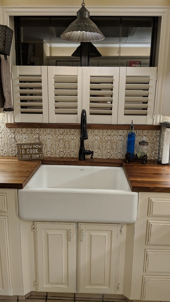 Photo of a small country u-shaped kitchen pantry in Dallas with a farmhouse sink, raised-panel cabinets, beige cabinets, wood benchtops, beige splashback, ceramic splashback, white appliances, ceramic floors, a peninsula, beige floor and brown benchtop.