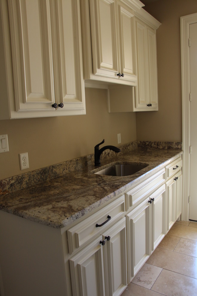 This is an example of a mid-sized mediterranean laundry room in Dallas with a single-bowl sink, raised-panel cabinets, white cabinets, granite benchtops, beige walls, travertine floors and a side-by-side washer and dryer.