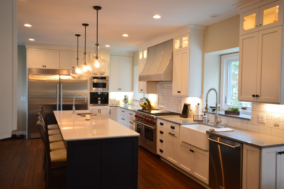 Photo of a large contemporary l-shaped open plan kitchen in Detroit with a farmhouse sink, shaker cabinets, white cabinets, quartz benchtops, white splashback, cement tile splashback, stainless steel appliances, bamboo floors, with island and brown floor.
