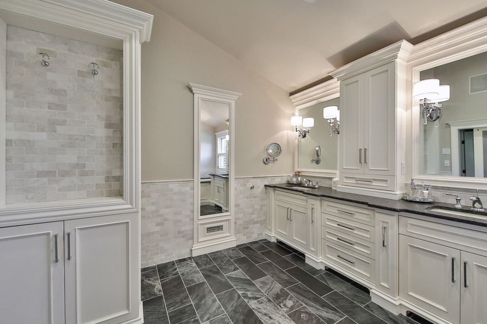 This is an example of a large transitional master bathroom in Other with beaded inset cabinets, white cabinets, an undermount tub, a corner shower, gray tile, marble, beige walls, an undermount sink, granite benchtops, grey floor, a hinged shower door and black benchtops.