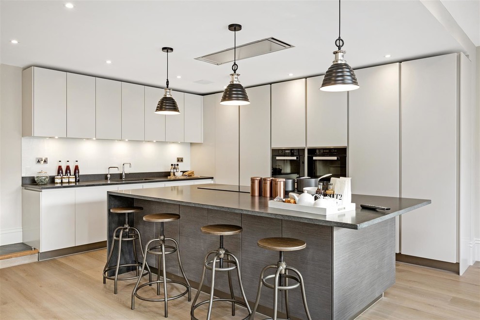 Mid-sized contemporary l-shaped kitchen in London with an undermount sink, flat-panel cabinets, white splashback, stainless steel appliances, with island, beige floor and black benchtop.