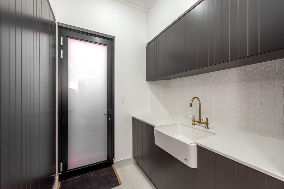 Photo of a contemporary galley dedicated laundry room in Perth with a farmhouse sink, black cabinets, solid surface benchtops, white splashback, mosaic tile splashback, white walls, porcelain floors, a stacked washer and dryer, grey floor and white benchtop.