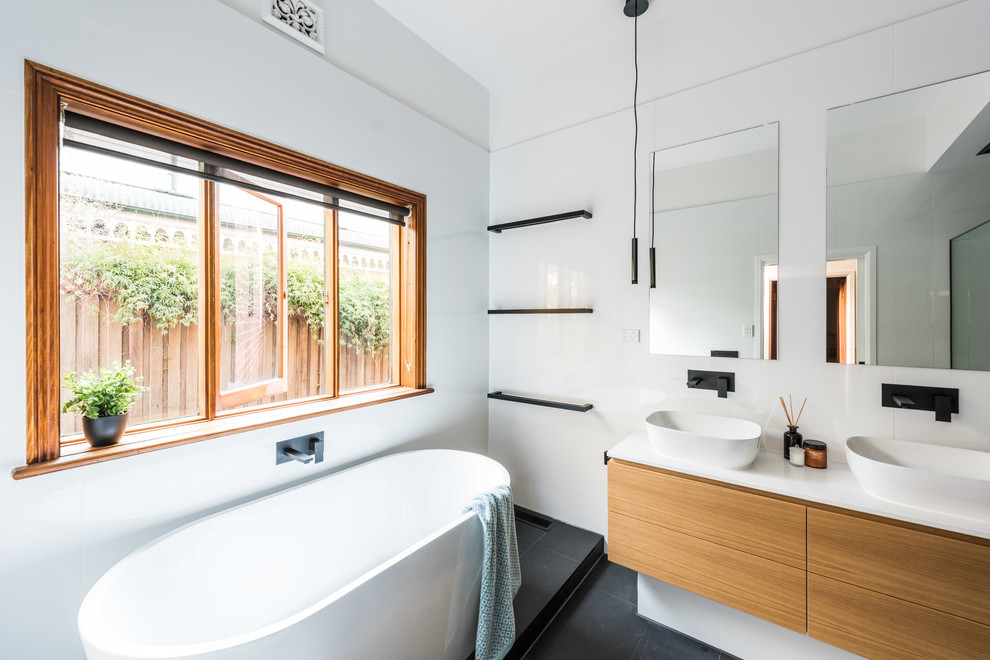 This is an example of a mid-sized modern master bathroom in Melbourne with flat-panel cabinets, a freestanding tub, white tile, ceramic tile, white walls, a vessel sink, engineered quartz benchtops, black floor, medium wood cabinets, a corner shower, a hinged shower door and white benchtops.