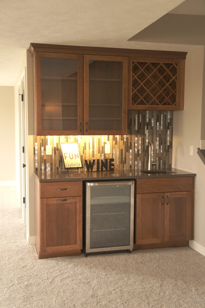 Photo of a small traditional single-wall wet bar in Omaha with an undermount sink, glass-front cabinets, medium wood cabinets, quartz benchtops, multi-coloured splashback, matchstick tile splashback, carpet, beige floor and brown benchtop.