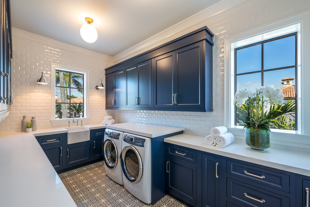 Photo of a beach style u-shaped dedicated laundry room in Miami with a farmhouse sink, recessed-panel cabinets, blue cabinets, a side-by-side washer and dryer, multi-coloured floor and white benchtop.