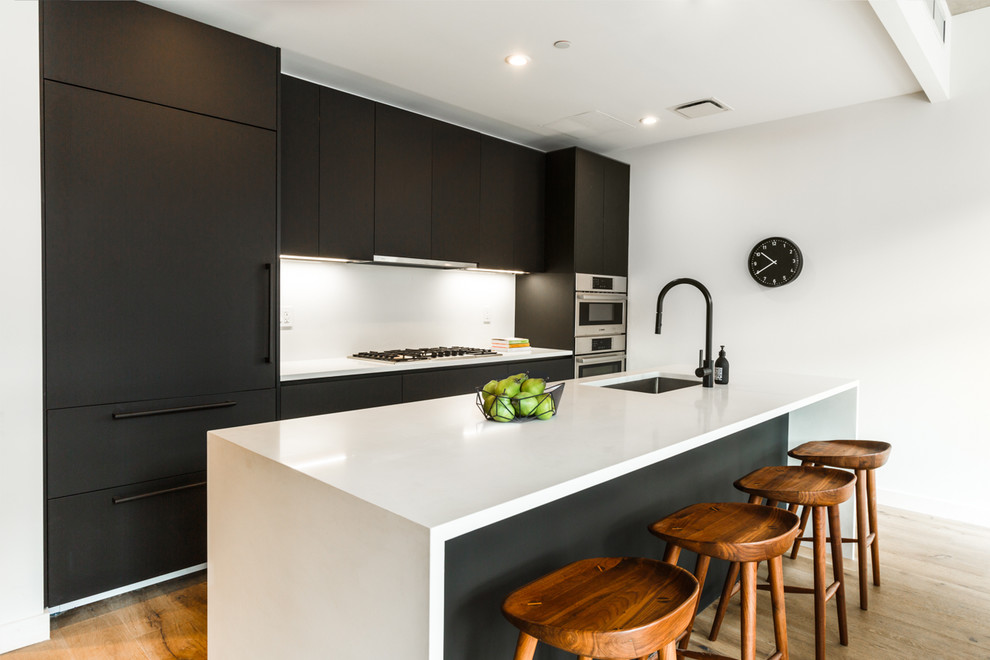 Inspiration for a mid-sized modern single-wall open plan kitchen in New York with an undermount sink, flat-panel cabinets, black cabinets, quartz benchtops, white splashback, stone slab splashback, panelled appliances, medium hardwood floors, with island, brown floor and white benchtop.