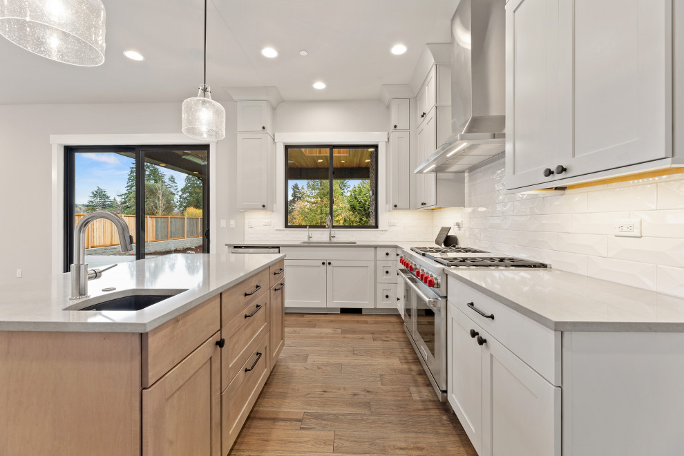 Inspiration for a mid-sized transitional l-shaped eat-in kitchen in Seattle with a drop-in sink, recessed-panel cabinets, white cabinets, quartz benchtops, white splashback, ceramic splashback, stainless steel appliances, medium hardwood floors, with island, brown floor and grey benchtop.