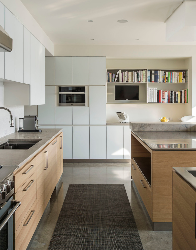 This is an example of a mid-sized modern galley kitchen in New York with an undermount sink, flat-panel cabinets, light wood cabinets, stainless steel benchtops, white splashback, stone tile splashback, stainless steel appliances, concrete floors and multiple islands.