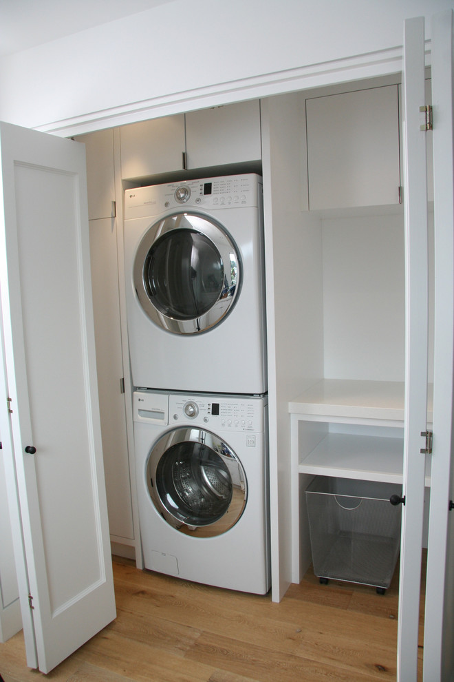 Mid-sized modern laundry room in San Francisco with flat-panel cabinets, white cabinets, solid surface benchtops, white walls, light hardwood floors and a stacked washer and dryer.