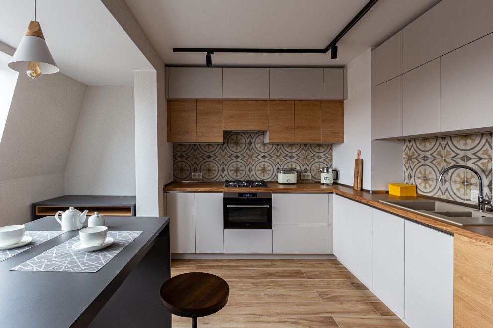 This is an example of a mid-sized contemporary l-shaped kitchen in Saint Petersburg with flat-panel cabinets, wood benchtops, multi-coloured splashback, ceramic splashback, black appliances, laminate floors, brown floor, brown benchtop, a double-bowl sink and grey cabinets.