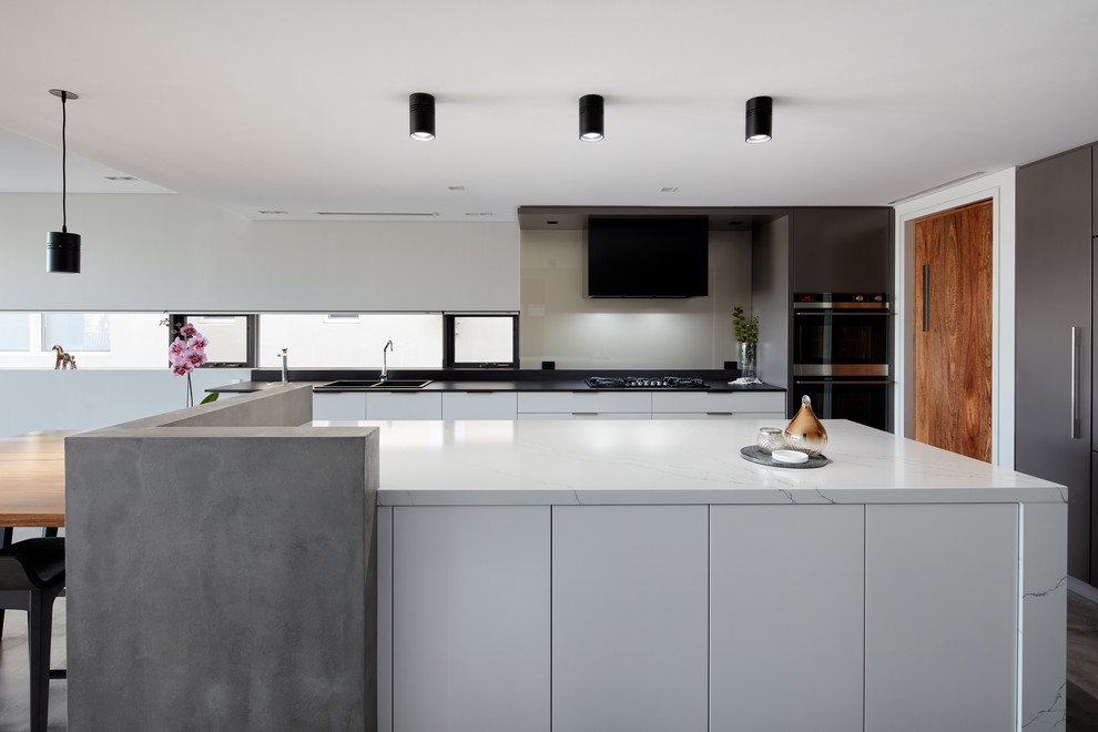 Photo of a contemporary galley eat-in kitchen in Sydney with a drop-in sink, flat-panel cabinets, white cabinets, grey splashback, glass sheet splashback, black appliances, with island, grey floor and white benchtop.