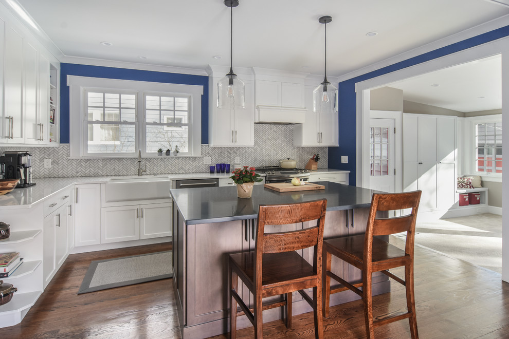 Mid-sized transitional l-shaped kitchen in New York with a farmhouse sink, shaker cabinets, white cabinets, quartz benchtops, white splashback, marble splashback, stainless steel appliances, dark hardwood floors, with island, brown floor and white benchtop.