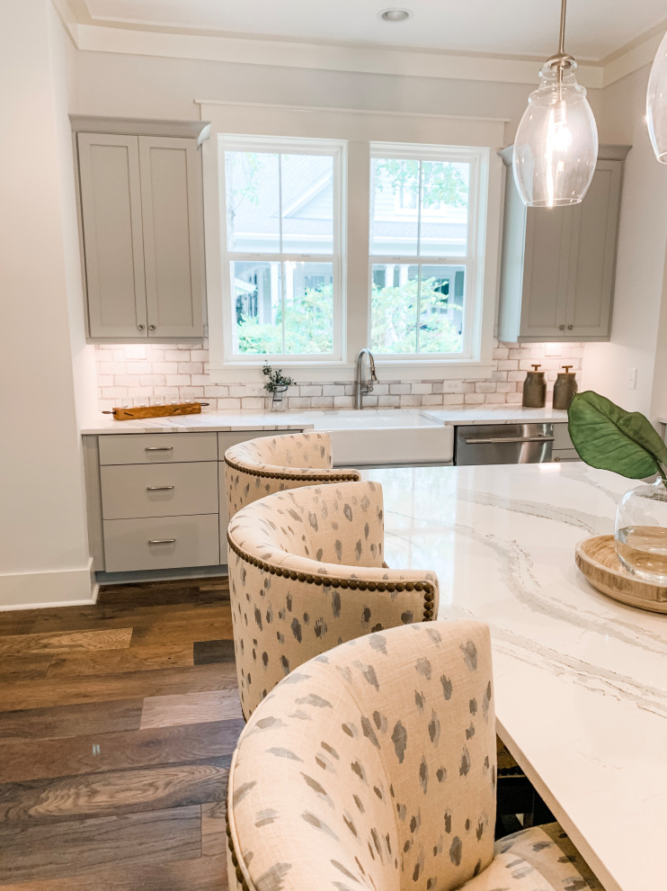 Mid-sized transitional l-shaped brown floor eat-in kitchen photo with a farmhouse sink, shaker cabinets, gray cabinets, quartz countertops, gray backsplash, ceramic backsplash, stainless steel appliances, an island and white countertops