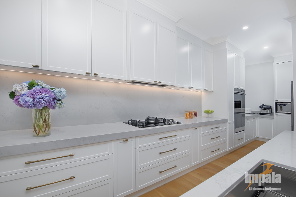Photo of a large galley open plan kitchen in Sydney with a submerged sink, shaker cabinets, engineered stone countertops, white splashback, engineered quartz splashback, stainless steel appliances, light hardwood flooring, an island and white worktops.