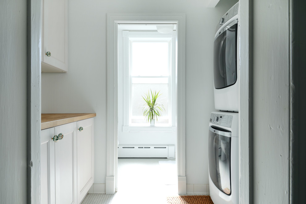 Photo of a mid-sized transitional galley dedicated laundry room in Philadelphia with recessed-panel cabinets, white cabinets, wood benchtops, white walls, porcelain floors, a stacked washer and dryer, white floor and brown benchtop.