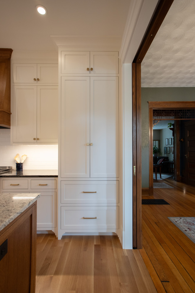 Large traditional u-shaped kitchen in Minneapolis with a farmhouse sink, recessed-panel cabinets, white cabinets, granite benchtops, white splashback, subway tile splashback, stainless steel appliances, medium hardwood floors, with island, brown floor and multi-coloured benchtop.