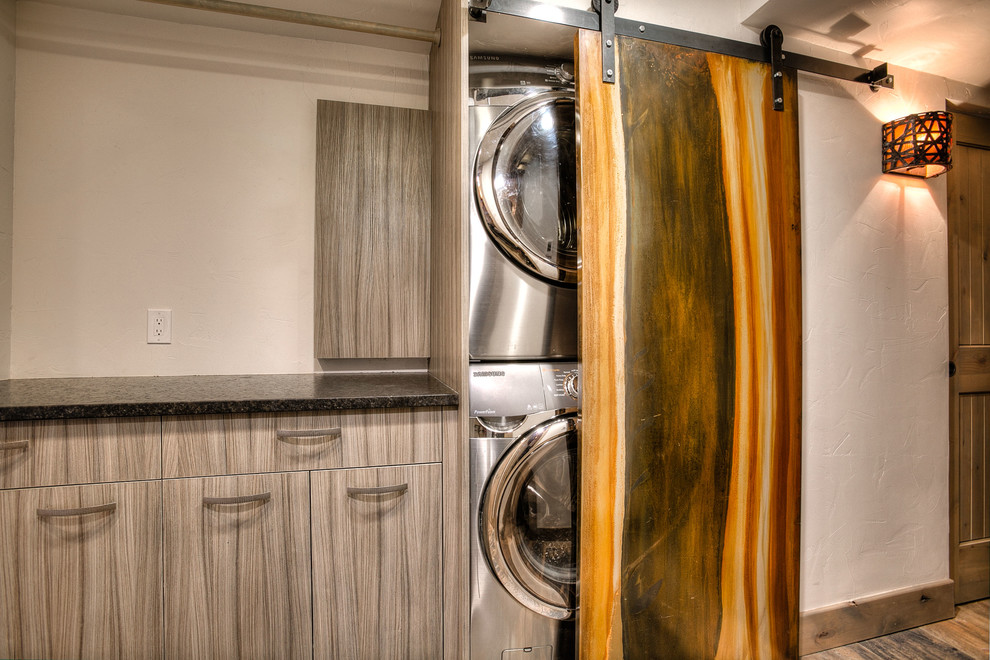 This is an example of a country single-wall laundry cupboard in Denver with flat-panel cabinets, grey cabinets, granite benchtops, grey walls, ceramic floors and a stacked washer and dryer.