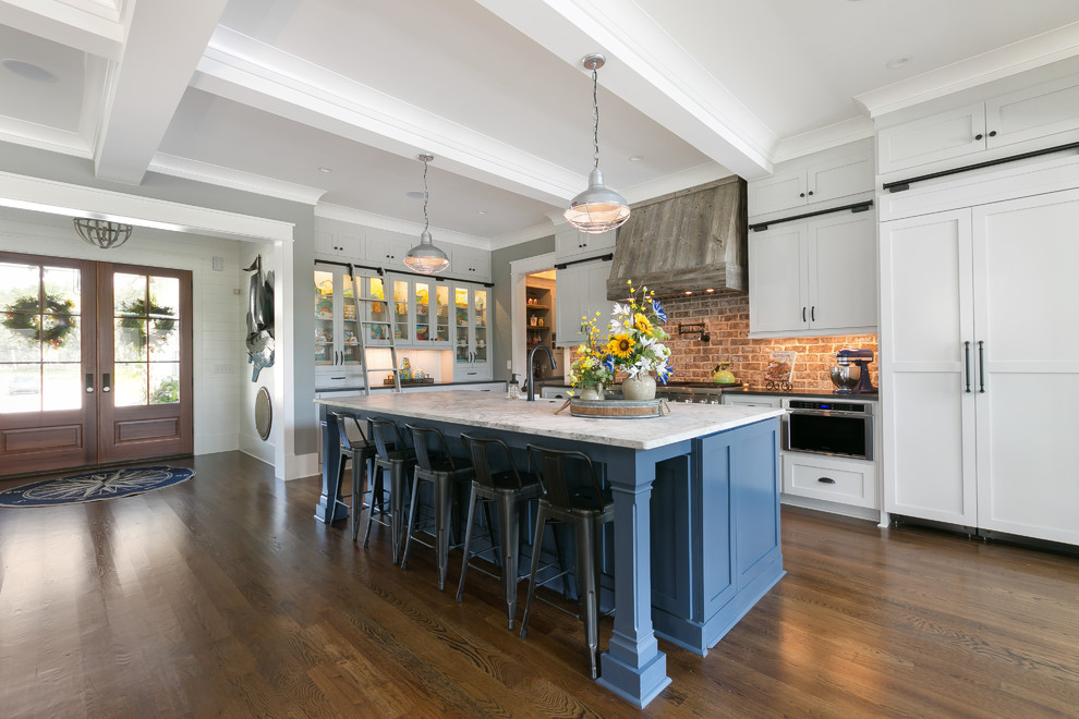 Photo of a beach style l-shaped kitchen in Charleston with a farmhouse sink, shaker cabinets, white cabinets, red splashback, brick splashback, stainless steel appliances, dark hardwood floors, with island, brown floor and white benchtop.