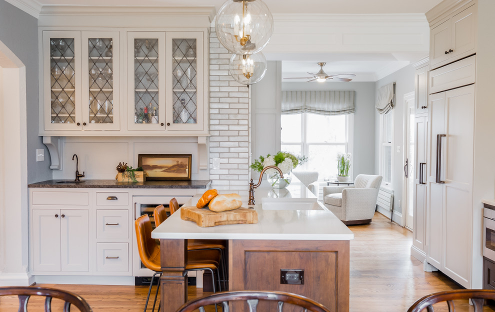 Large transitional u-shaped open plan kitchen in St Louis with a farmhouse sink, recessed-panel cabinets, white cabinets, quartz benchtops, white splashback, porcelain splashback, panelled appliances, medium hardwood floors, a peninsula, brown floor and white benchtop.