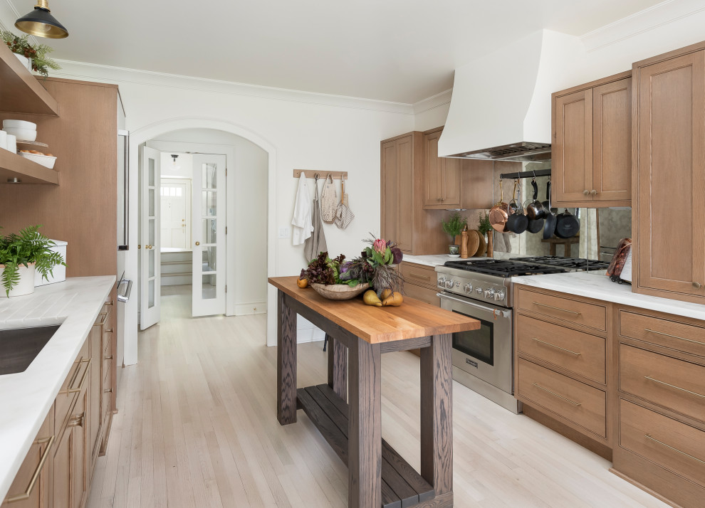 Photo of a mid-sized transitional galley kitchen in Chicago with beaded inset cabinets, medium wood cabinets, marble benchtops, metallic splashback, mirror splashback, stainless steel appliances, light hardwood floors, with island, white benchtop, an undermount sink and beige floor.