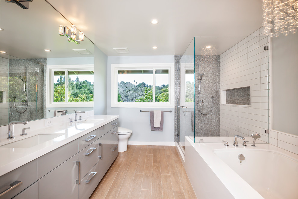 This is an example of a large contemporary master bathroom in San Francisco with flat-panel cabinets, grey cabinets, an undermount tub, a corner shower, gray tile, white tile, grey walls, an undermount sink, a two-piece toilet, ceramic tile, porcelain floors, brown floor, a hinged shower door and white benchtops.